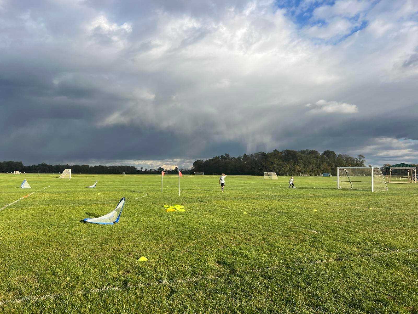 A youth soccer field under dark storm clouds at the end of a tournament day, a couple of kids still out on the grass among portable goals and cones