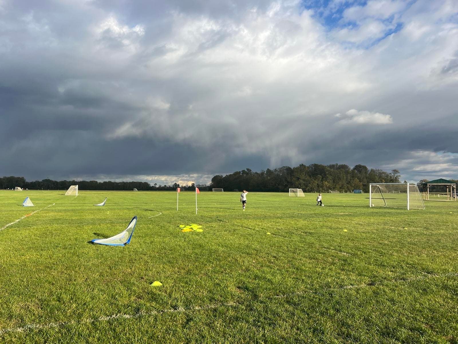 A youth soccer field under dark storm clouds at the end of a tournament day, a couple of kids still out on the grass among portable goals and cones